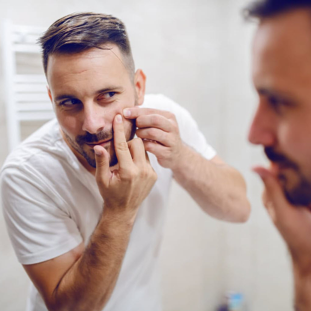 Man in a white t-shirt looking at his reflection in a mirror, touching his face.